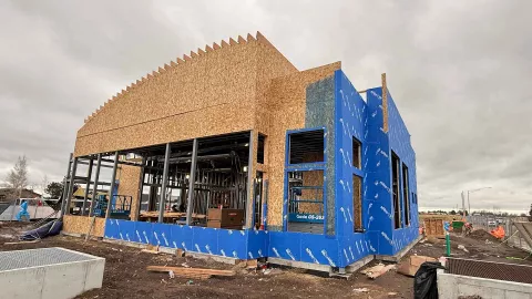 AFCU Rexburg branch wood framing in progress — full exterior view showing the building's curved arc roof feature, mixed wood-and-steel structural system, and blue weather-resistant barrier