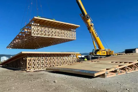 Mountain Crane lifting a prefabricated wood truss section into place at the Alpine Hotel site, with a BLU-D crew member spotting from the ground
