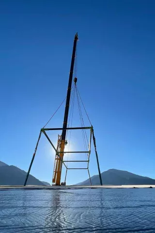 Backlit silhouette of a crane and spreader-bar rigging staged on the Alpine Hotel deck, framed against bright sun and mountain peaks