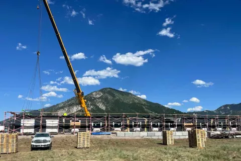 Wide shot of the Alpine Hotel under construction with scaffolding wrapped around the structure, a yellow boom crane on site, and mountains in the background