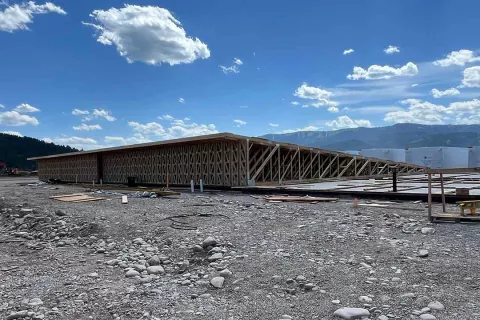 Truss-framed wall section at the Alpine Hotel viewed across the river-rock construction yard, with mountains and scattered clouds beyond