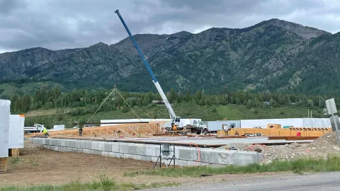 Mobile crane lifting a wood truss at the Alpine Hotel project site, with framers at work on the foundation slab and the Snake River Range rising behind