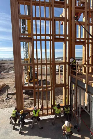 BLU-D crew of six in high-vis hoisting a tall, multi-story wood-framed wall section into position at Grand Peaks, with a telehandler staged behind