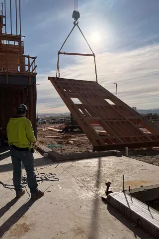 Crane-suspended wood-framed wall panel backlit by the sun above the Grand Peaks slab, with a tagline-rope spotter watching from the foreground