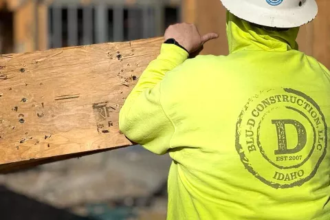 Crew member in a BLU-D Construction high-vis hoodie carrying a piece of OSB sheathing, the company's Est. 2007 Idaho logo visible across the back