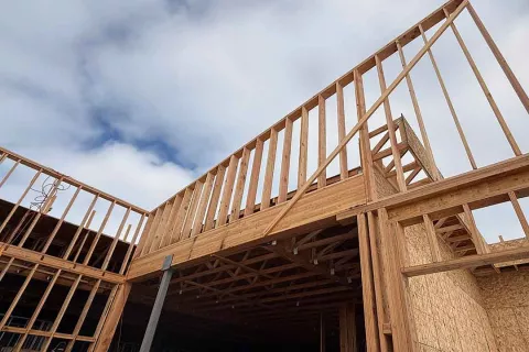Looking up at the Grand Peaks framed roof structure where wood-framed walls and trusses meet a steel column under a partly cloudy sky