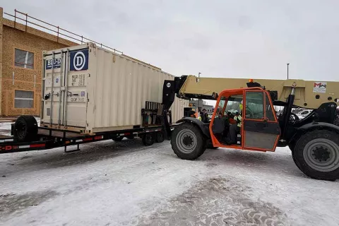 Telehandler positioning a BLU-D Construction job-site shipping container on a flatbed trailer at the snowy Grand Peaks site