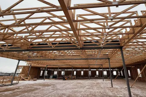 Interior view at Grand Peaks showing engineered roof trusses spanning above steel beams and columns, with the floor below dusted with snow