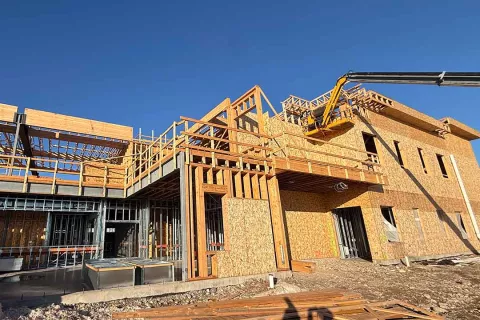 Interior of the Grand Peaks shell with sunlight streaming through wood-framed walls and trusses, steel beams running across, and a framer at work on temporary scaffolding to the left