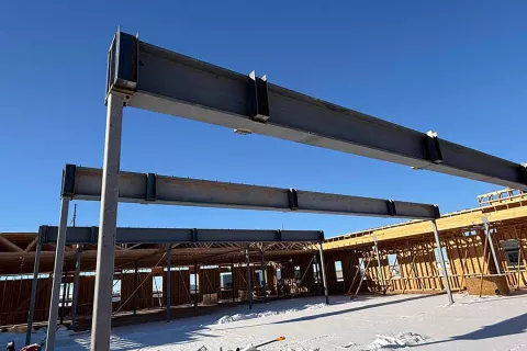 Structural steel beams spanning between columns at Grand Peaks under a clear blue sky, with snow on the concrete slab and wood-framed walls beyond