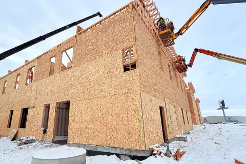 Two-story OSB-sheathed exterior wall at Grand Peaks with two articulated boom lifts positioning framers at the upper roof line, snow on the ground