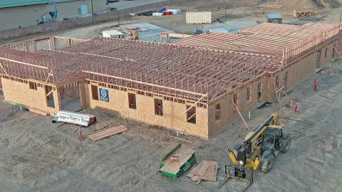 Aerial view of the IDOC Re-Entry Center under construction with OSB-sheathed wood-framed walls, engineered roof trusses spanning the building, a yellow CAT telehandler staged at the foreground, and a BLU-D logo banner mounted on the side