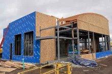 AFCU Rexburg branch under construction — wood framing on blue WRB-wrapped exterior, steel structural frame, and the building's signature curved arc roof feature taking shape