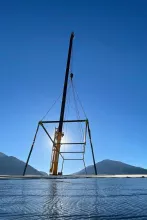 Backlit silhouette of a crane and spreader-bar rigging staged on the Alpine Hotel deck, framed against bright sun and mountain peaks