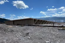 Truss-framed wall section at the Alpine Hotel viewed across the river-rock construction yard, with mountains and scattered clouds beyond