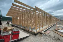 Long wood-framed wall lying flat on the Alpine Hotel foundation with a framer at work, Simpson hardware boxes and Milwaukee tool cases staged beside it under stormy skies