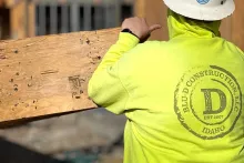 Crew member in a BLU-D Construction high-vis hoodie carrying a piece of OSB sheathing, the company's Est. 2007 Idaho logo visible across the back