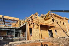 Interior of the Grand Peaks shell with sunlight streaming through wood-framed walls and trusses, steel beams running across, and a framer at work on temporary scaffolding to the left