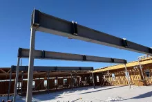 Structural steel beams spanning between columns at Grand Peaks under a clear blue sky, with snow on the concrete slab and wood-framed walls beyond