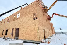 Two-story OSB-sheathed exterior wall at Grand Peaks with two articulated boom lifts positioning framers at the upper roof line, snow on the ground