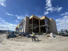 Alpine Academy exterior under construction, the masonry and steel shell with wood framing showing through the glazed stair tower, a boom lift at work