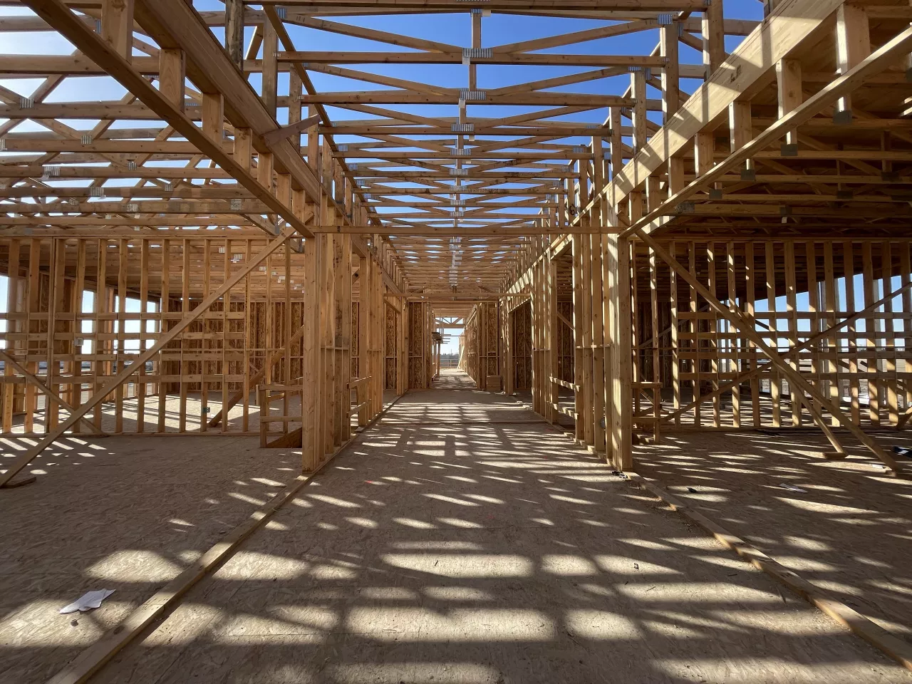 Long second-floor corridor at Alpine Academy, wood-framed classroom walls on both sides under open roof framing, sunlight casting a grid of shadows on the subfloor