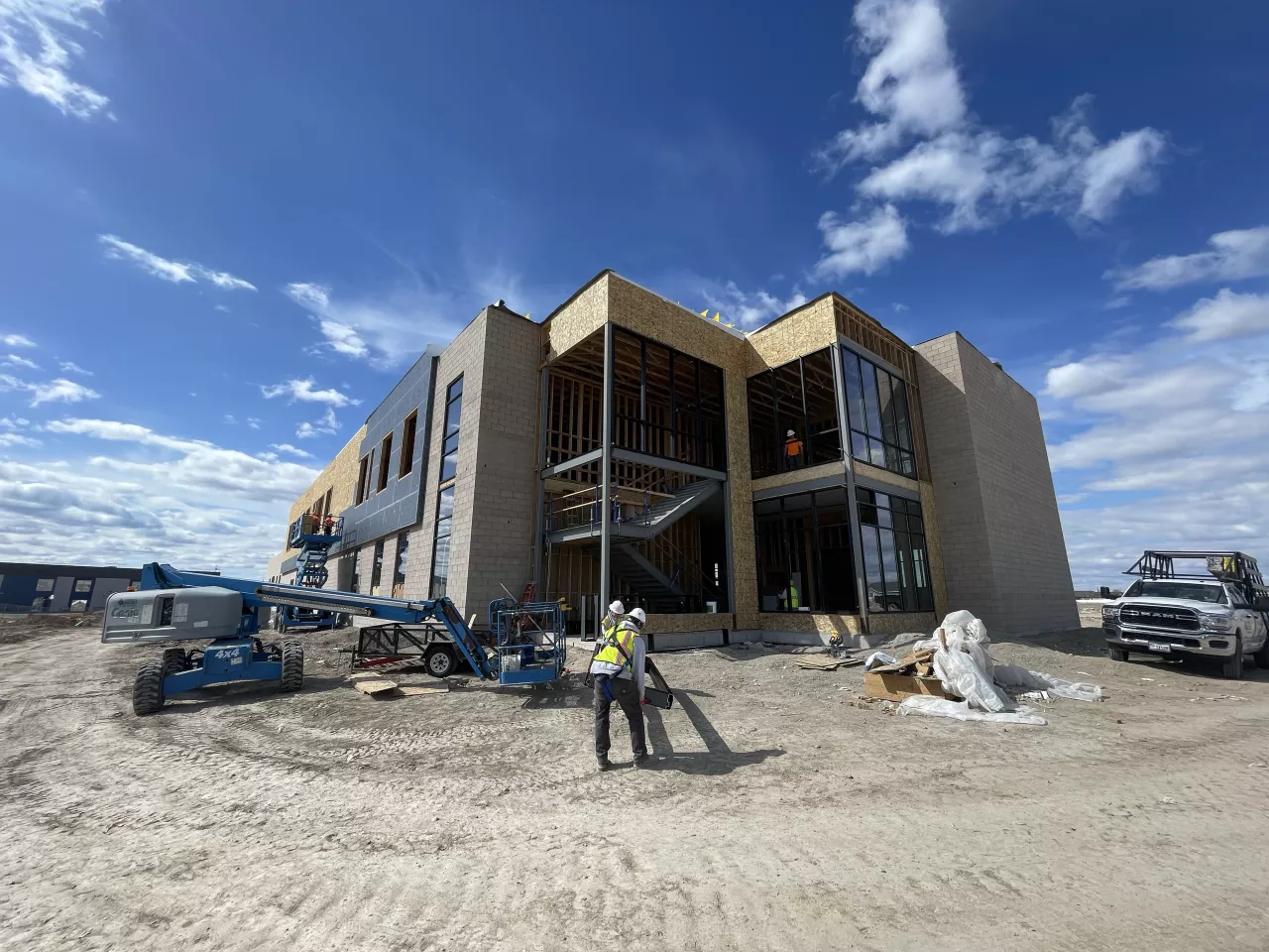 Alpine Academy exterior under construction, the masonry and steel shell with wood framing showing through the glazed stair tower, a boom lift at work