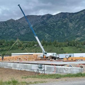 Mobile crane lifting a wood truss at the Alpine Hotel project site, with framers at work on the foundation slab and the Snake River Range rising behind