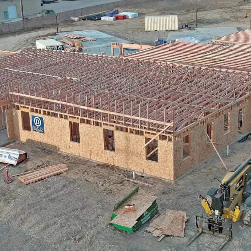 Aerial view of the IDOC Re-Entry Center under construction with OSB-sheathed wood-framed walls, engineered roof trusses spanning the building, a yellow CAT telehandler staged at the foreground, and a BLU-D logo banner mounted on the side